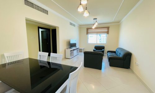 Modern apartment living room available for rent in Al Burhama, featuring a dining table with chairs, a couch, an armchair, and a TV unit, with natural light coming through a large window