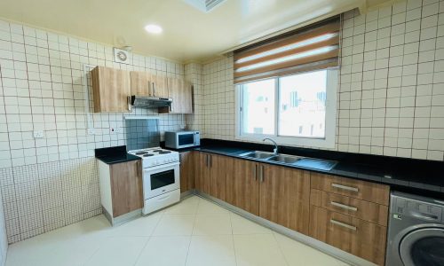 A modern kitchen with wooden cabinets, black countertops, and built-in appliances, including a microwave, stove, and washing machine, by a window in an Al Burhama apartment for rent.