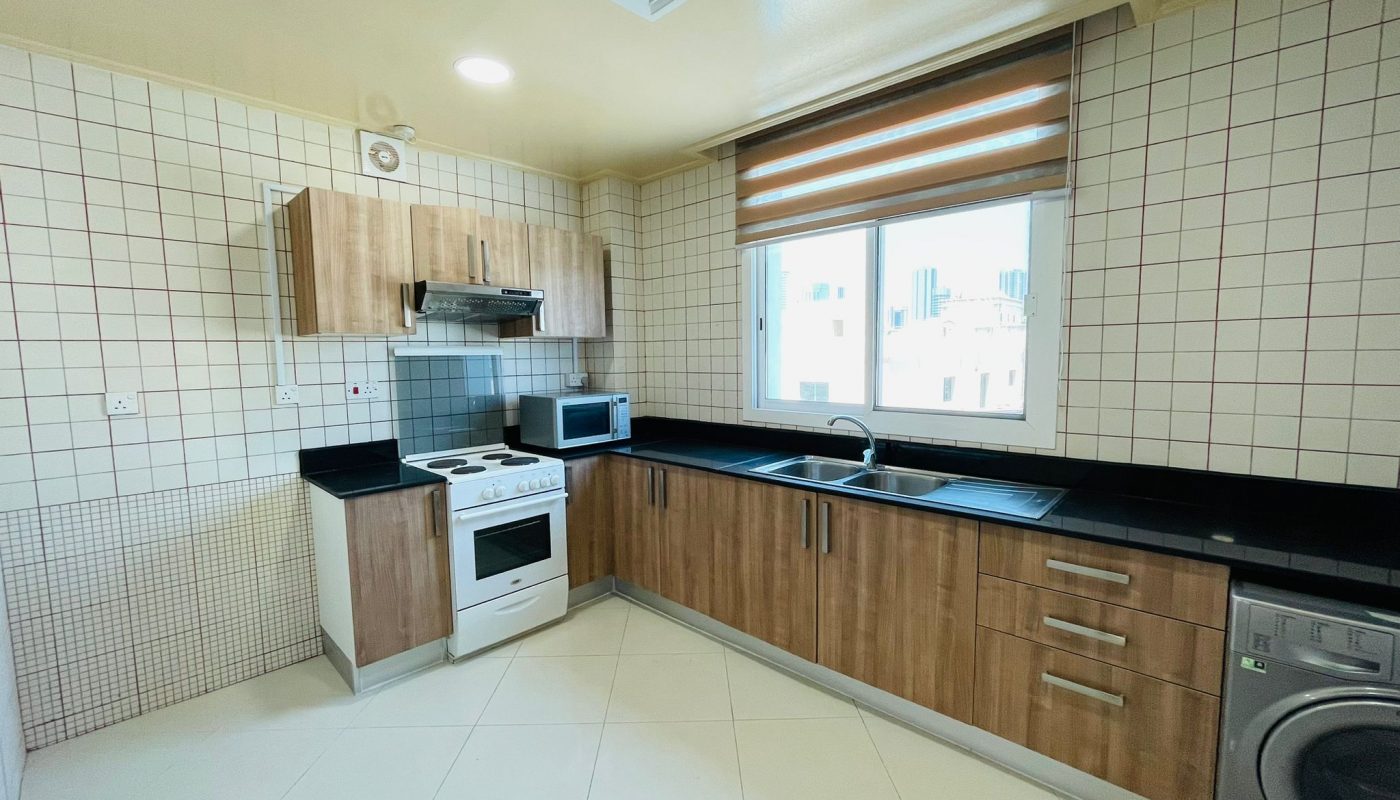 A modern kitchen with wooden cabinets, black countertops, and built-in appliances, including a microwave, stove, and washing machine, by a window in an Al Burhama apartment for rent.