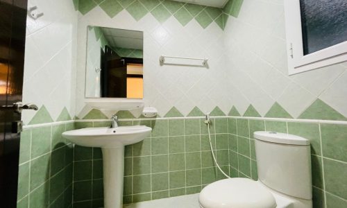 A clean bathroom in an apartment for rent, featuring a white sink, toilet, and a mirror on a wall tiled with pale green tiles. A window is visible on the left.