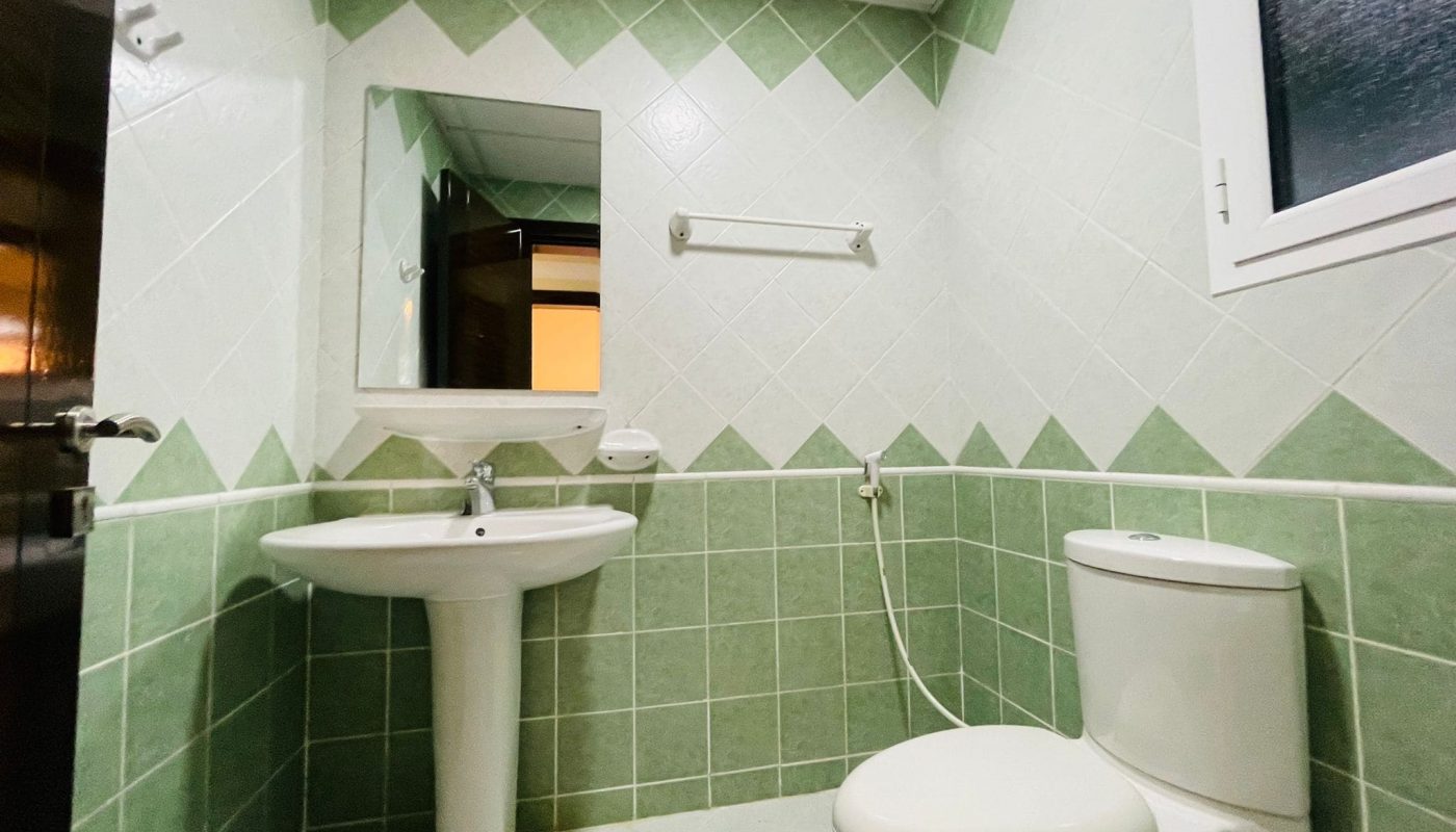 A clean bathroom in an apartment for rent, featuring a white sink, toilet, and a mirror on a wall tiled with pale green tiles. A window is visible on the left.