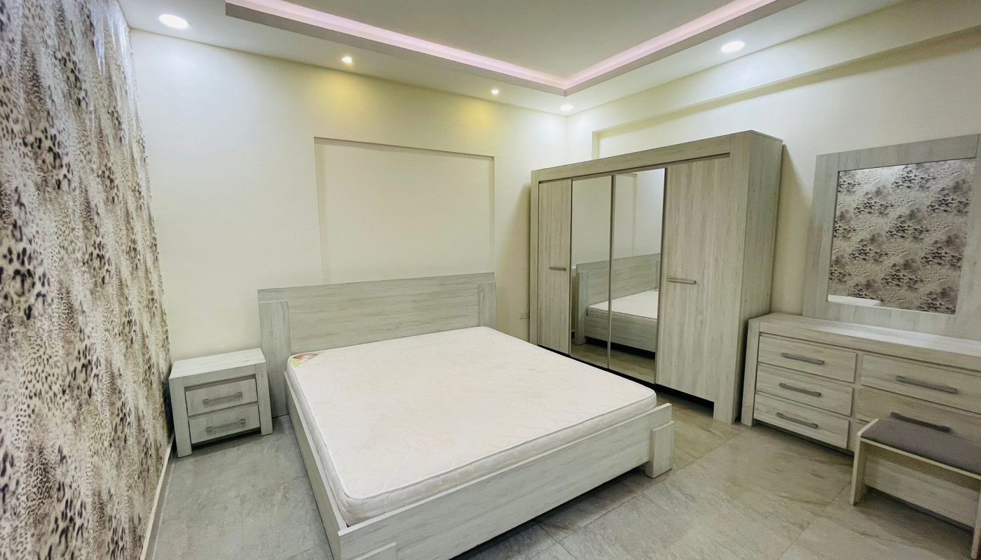 Modern studio interior in Al Burhama with a double bed, built-in wardrobes, and ambient lighting. Decor includes textured wall panels and a full-length mirror.