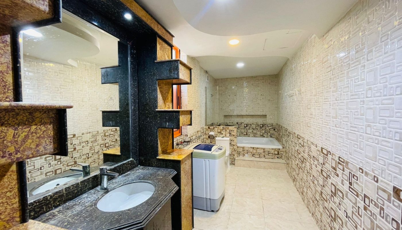 Modern studio apartment bathroom in Al Burhama, featuring geometric tiled walls, a white ceramic sink, rectangular mirror, and a washing machine.