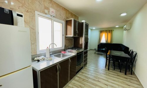 A compact kitchen with wooden cabinets, a built-in stove, and a sink in an available apartment, leading to a dining area with a black table and chairs.