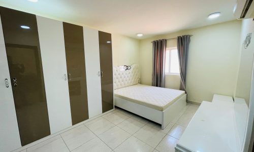 A small, well-lit bedroom in an available apartment features a single bed, large wardrobe with brown and white doors, and a window with draped curtains.