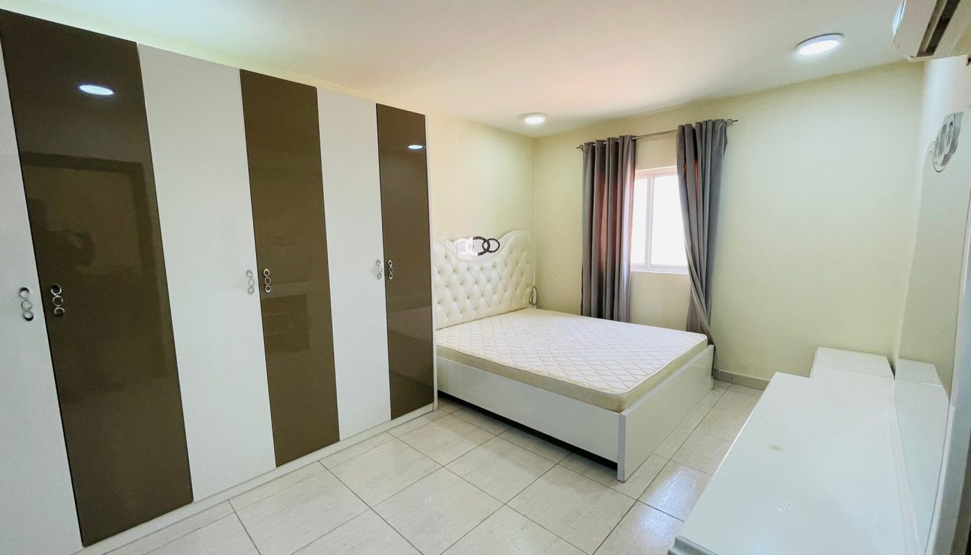 A small, well-lit bedroom in an available apartment features a single bed, large wardrobe with brown and white doors, and a window with draped curtains.