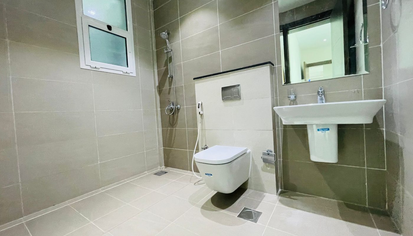 Modern bathroom interior with beige tiles in an Al Burhama apartment, featuring a wall-mounted sink, toilet, shower area, and a mirror. A small window provides natural light.
