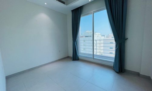 Empty modern apartment for rent with white walls and a large window covered by drawn teal curtains, showing a building outside in Al Burhama.