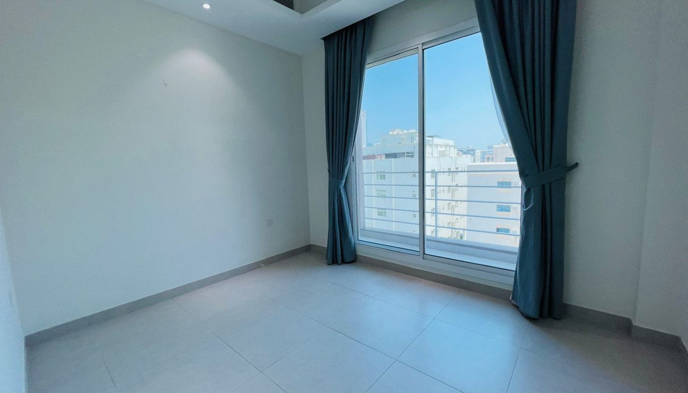 Empty modern apartment for rent with white walls and a large window covered by drawn teal curtains, showing a building outside in Al Burhama.
