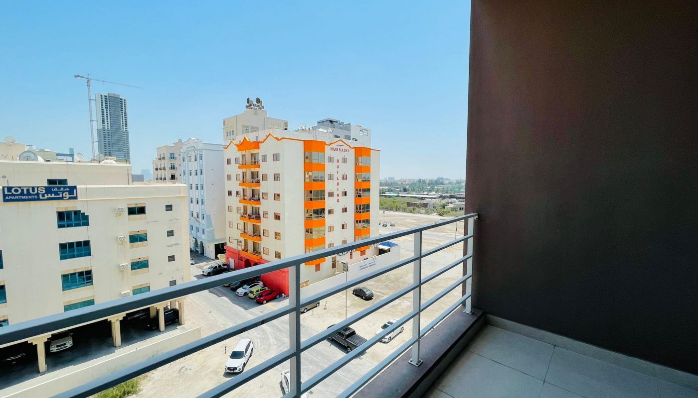 View from an available apartment balcony overlooking a bright sunny cityscape with modern buildings, clear skies, and a narrow road.