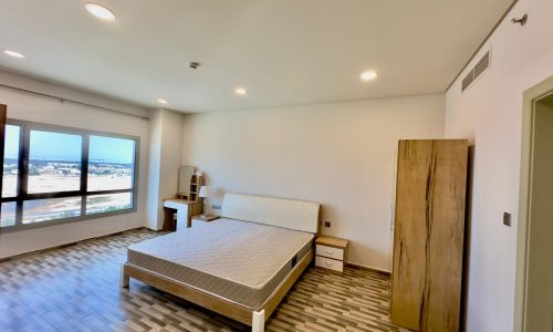 A luxury apartment bedroom with a large bed, herringbone floor, wardrobe, and ceiling lights, featuring a wide window with a city view.