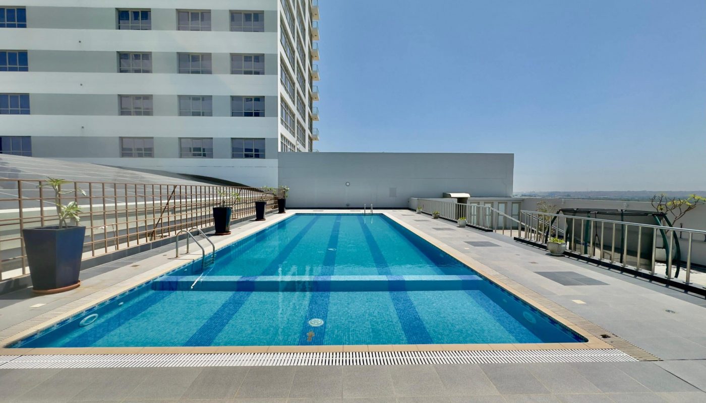 Rooftop swimming pool on a sunny day, flanked by a luxury apartment building and clear blue sky, with a cityscape visible in the distance.
