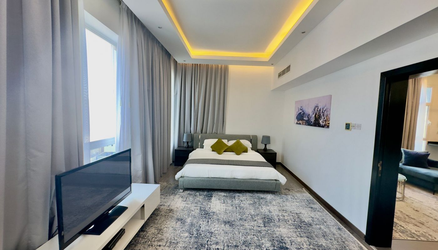 Luxury apartment room with a large bed, gray curtains, ambient lighting, carpeted floor, and a flat-screen TV.