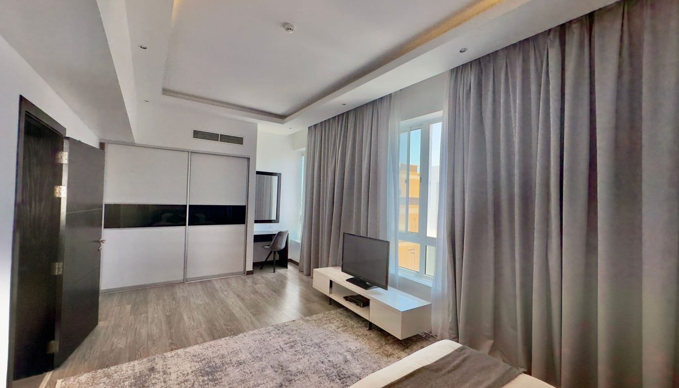 Luxury apartment with a large bed, gray curtains, hardwood flooring, a TV, and bright natural light from a window.