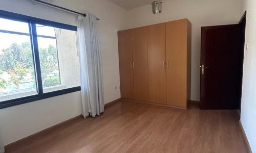 Empty room with wooden flooring, an auto draft, a large brown wardrobe, white curtains, and a door on the right. A window offers a view of trees outside.