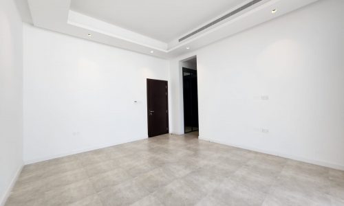 A spacious, empty room in a luxury villa with white walls and grey tiled flooring, featuring recessed lighting and a dark wooden door.