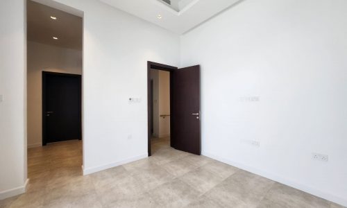 Empty room within a private villa in Saar, featuring white walls, gray tiled floor, and two dark wooden doors; one open and one closed.