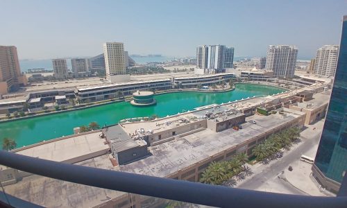 Aerial view of a modern waterfront development in Amwaj with residential buildings, palm trees, and a circular structure on the pier, viewed from a balcony.