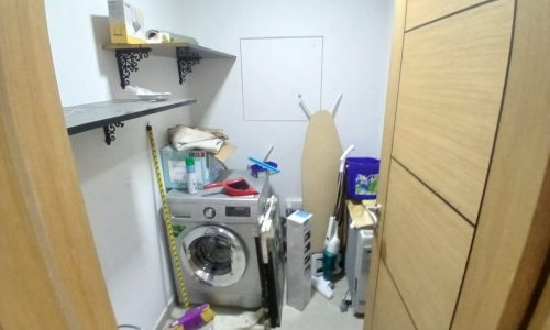 A cluttered utility room in a modern flat with a washing machine, ironing board, and various household items stored on shelves and the floor.