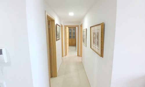 A brightly lit hallway in a modern flat, featuring beige tiled floors, white walls, wooden doors, and framed paintings along the walls.