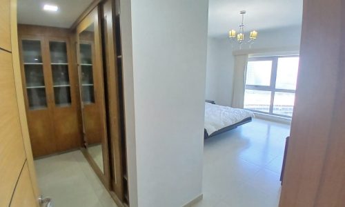 Interior of a modern flat showing an open wooden door leading to a bedroom with a bed and window, adjacent to a hallway with a wardrobe.