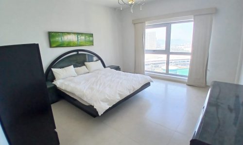 Modern flat with a black bed frame and white bedding, featuring a large window with a city view, sleek furnishings, and a hanging chandelier.