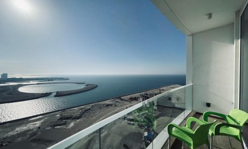 Balcony view overlooking a calm sea, a curved shoreline, and a clear blue sky, with a sunny glare and two green chairs visible in the amazing Studio.