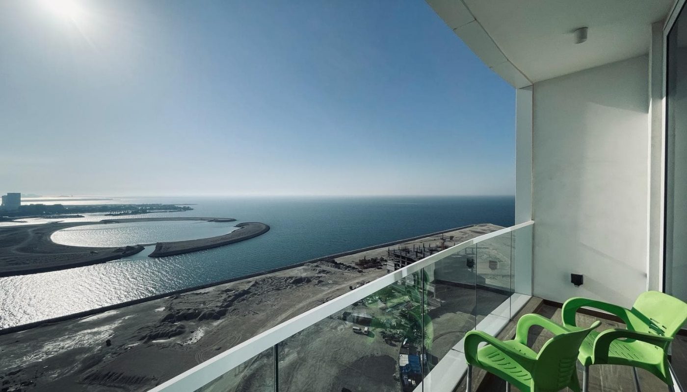 Balcony view overlooking a calm sea, a curved shoreline, and a clear blue sky, with a sunny glare and two green chairs visible in the amazing Studio.