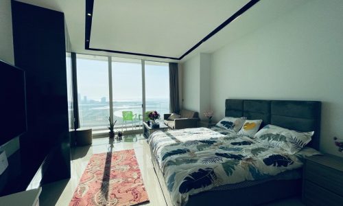 Studio for Rent: Modern bedroom with a large bed featuring palm leaf-patterned linens, a red rug, and floor-to-ceiling windows revealing an amazing city view in the Seef area.