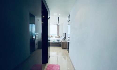 A modern apartment hallway leading to a bedroom with an ocean view in the Seef area, featuring a pink rug, wood flooring, and light walls.