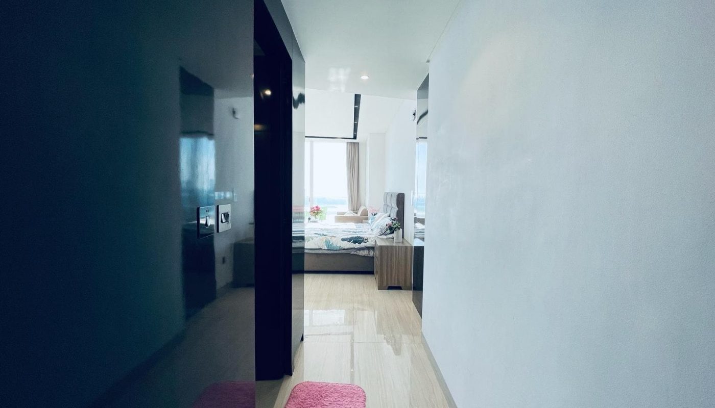 A modern apartment hallway leading to a bedroom with an ocean view in the Seef area, featuring a pink rug, wood flooring, and light walls.