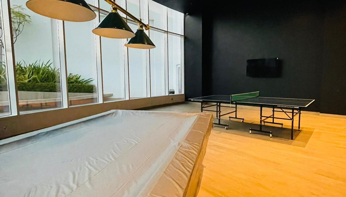 A room with a covered billiards table, a ping pong table, a wall-mounted TV, large windows, and modern ceiling lights.