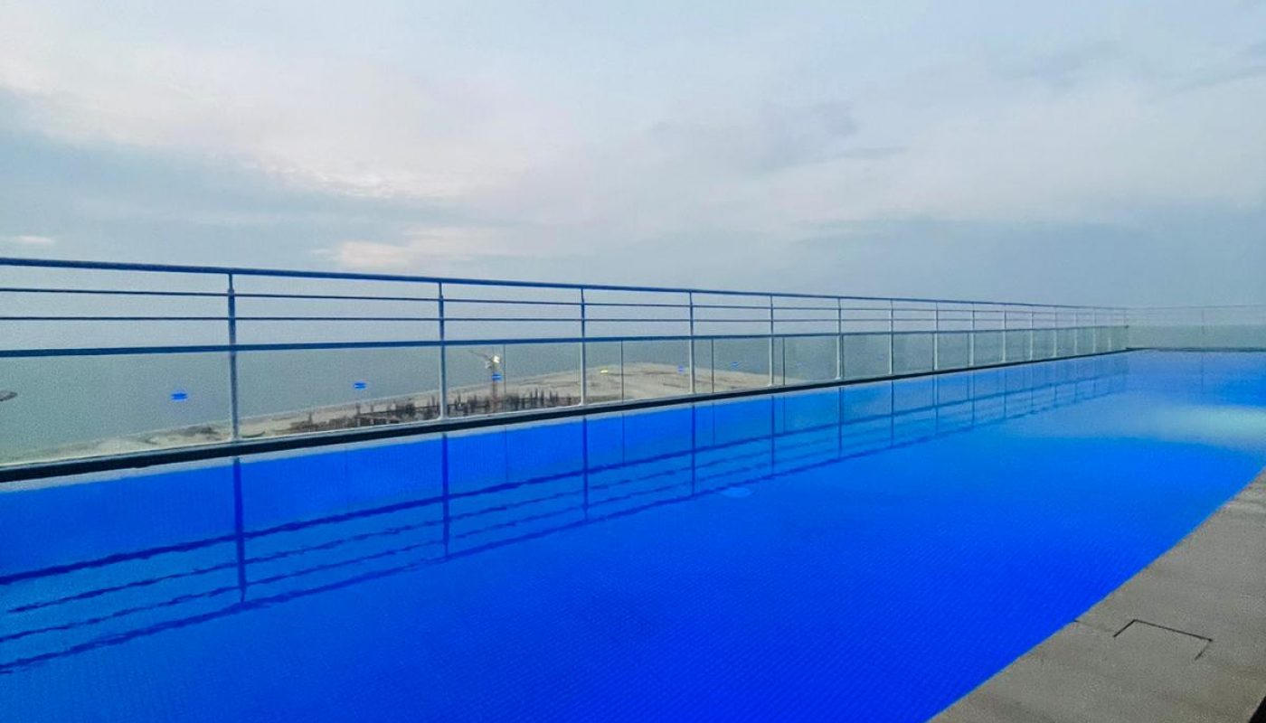 Infinity pool with blue water overlooking a calm ocean and cloudy sky, bordered by a wooden deck and metal railing.