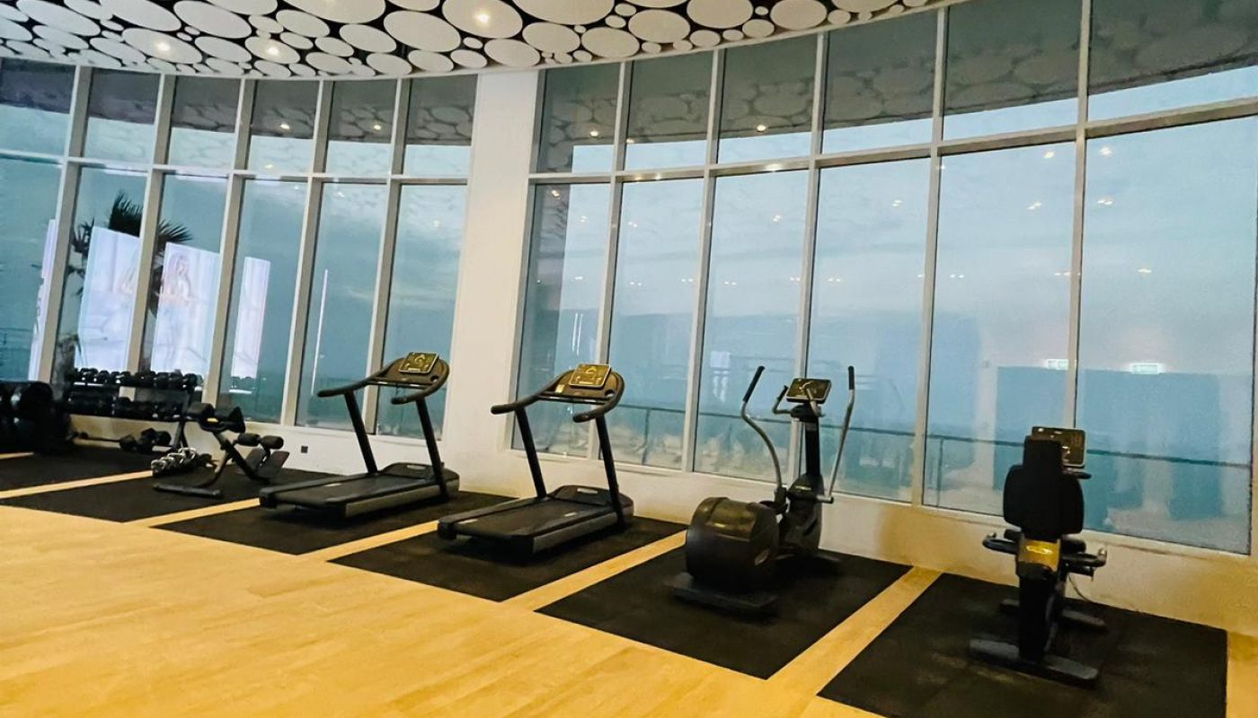 Modern gym with light wood flooring, black exercise equipment including treadmills and bikes, and large windows. The ceiling features a circular pattern design.