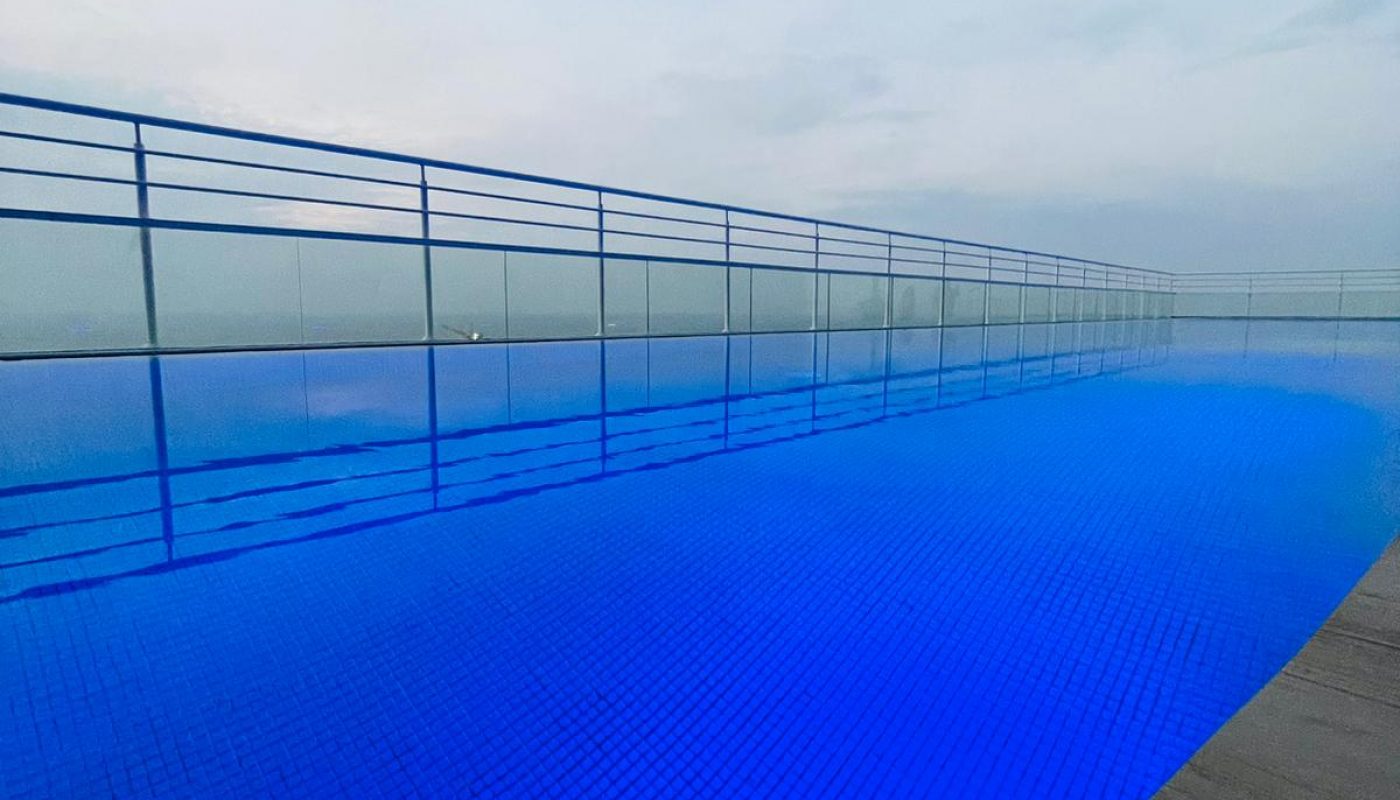 Infinity pool with blue water tiles, bordered by a wooden deck and metal railing, overlooking a hazy sky at dusk or dawn.