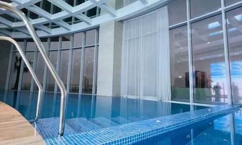 Indoor infinity pool with a tiled edge and metal handrails, surrounded by floor-to-ceiling windows and a ceiling with a grid pattern. White curtains partially cover some of the windows.