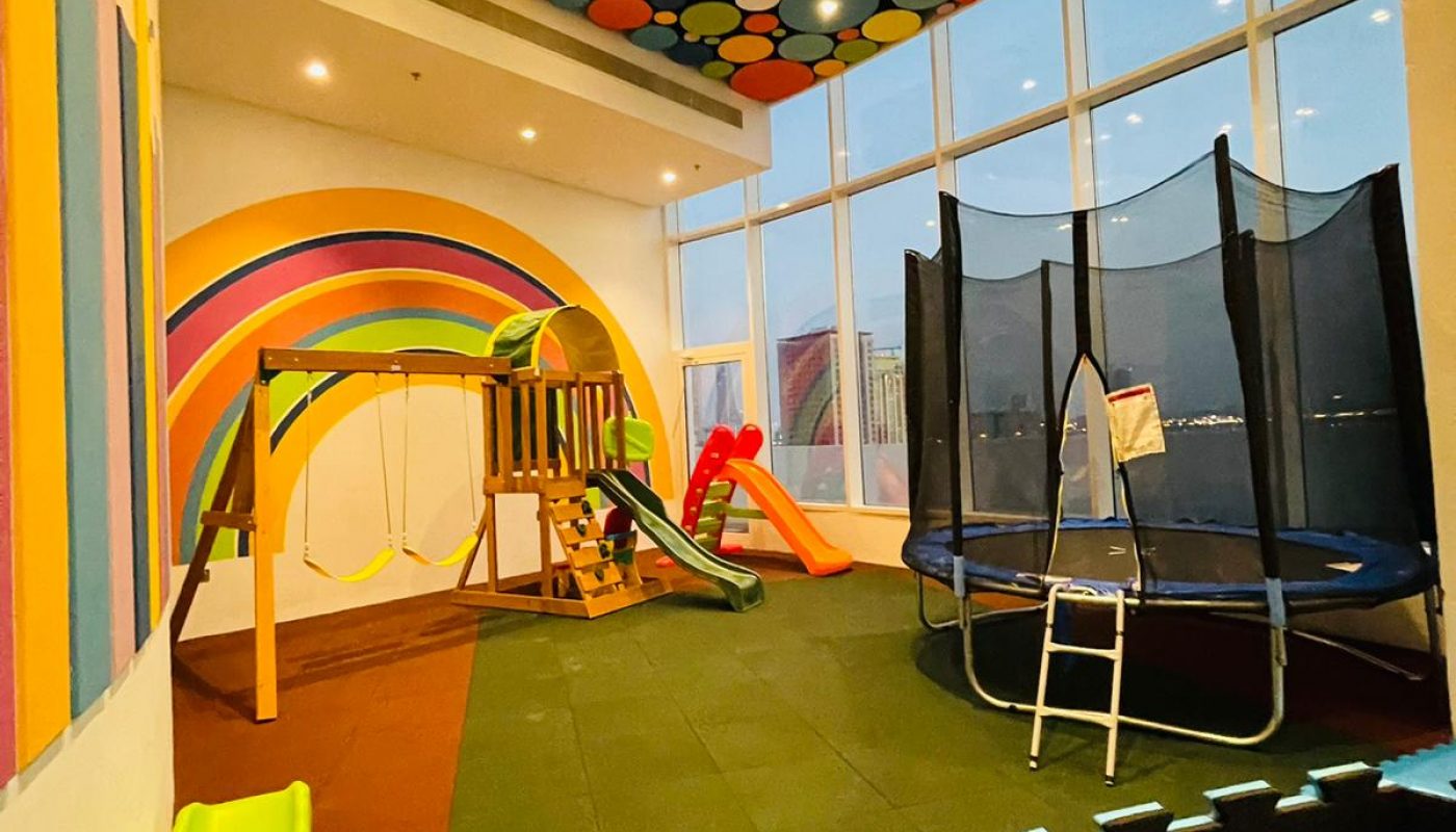 Indoor children's play area with colorful wall and ceiling designs, a slide, climbing structure, trampoline, and padded flooring. Large windows provide natural light.