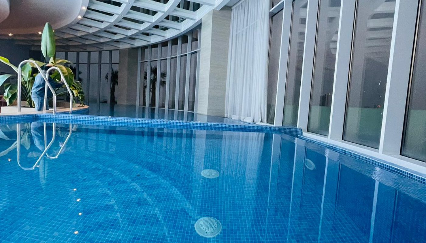 An indoor swimming pool with blue tiles, surrounded by floor-to-ceiling windows and a curved glass ceiling, with some plants in the corner.
