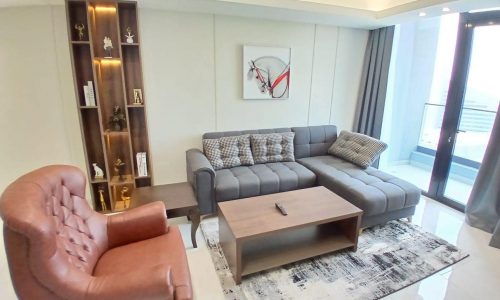 Modern living room with a large gray sofa, matching chair, coffee table, decorative shelf, and abstract wall art.