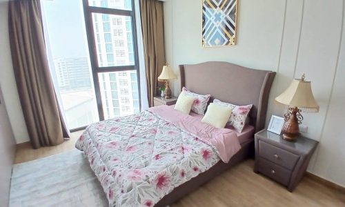 Modern bedroom in an Amwaj flat with large bed covered in floral bedding, two bedside tables with lamps, an artwork on the wall, and a large window with city view.