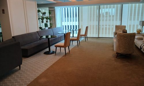 Modern office lounge area with large windows, a gray sofa, patterned armchair, and a round black Auto Draft table.