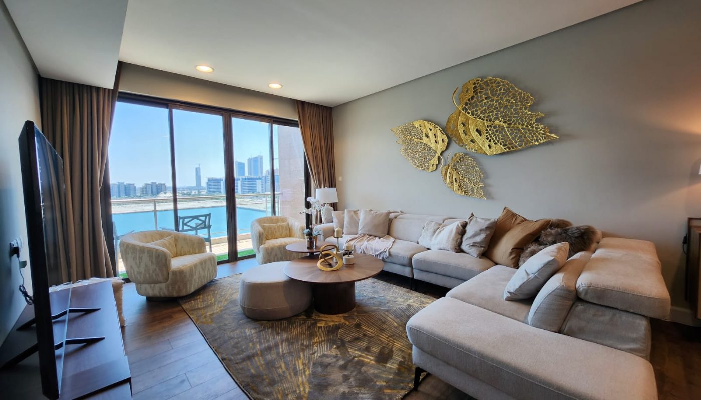 Modern living room with a large sectional sofa, coffee table, decorative wall art, floor lamp, and a city and waterfront view through large windows.