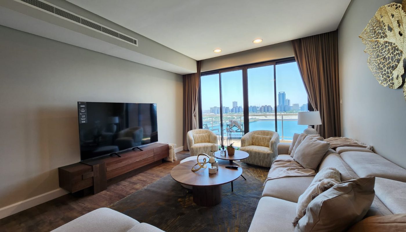 A modern living room with a large beige sectional sofa, a flat-screen TV, two armchairs, a round coffee table, and large windows with a city and waterfront view.