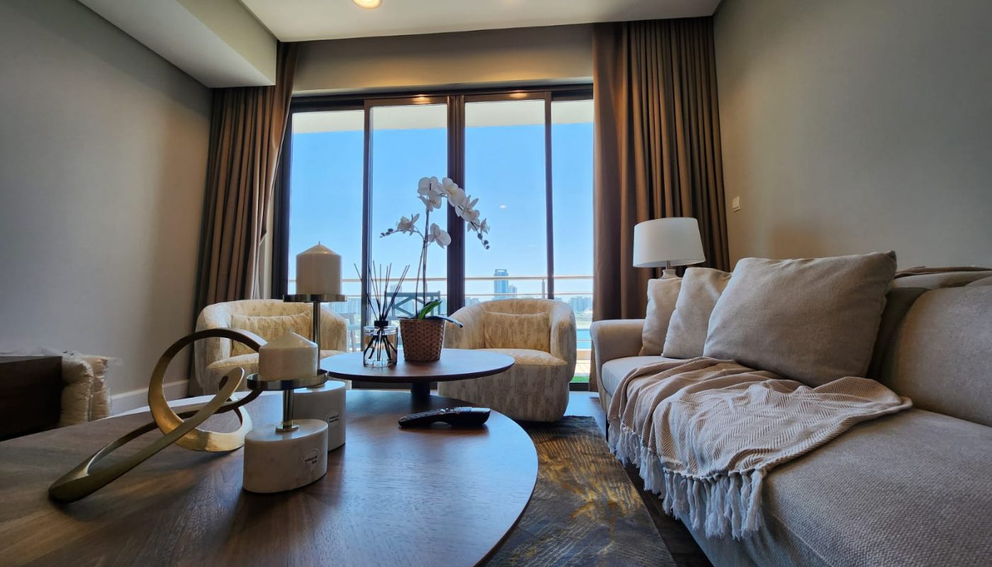 A modern living room with a beige sofa, coffee table with decor, large windows with brown curtains, and a view of a balcony and clear sky outside.