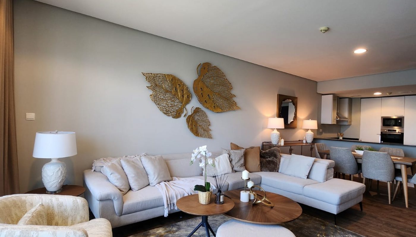 Modern living room with neutral-toned furniture, a round wooden coffee table, decorative wall art, and an open view of the dining area and kitchen.