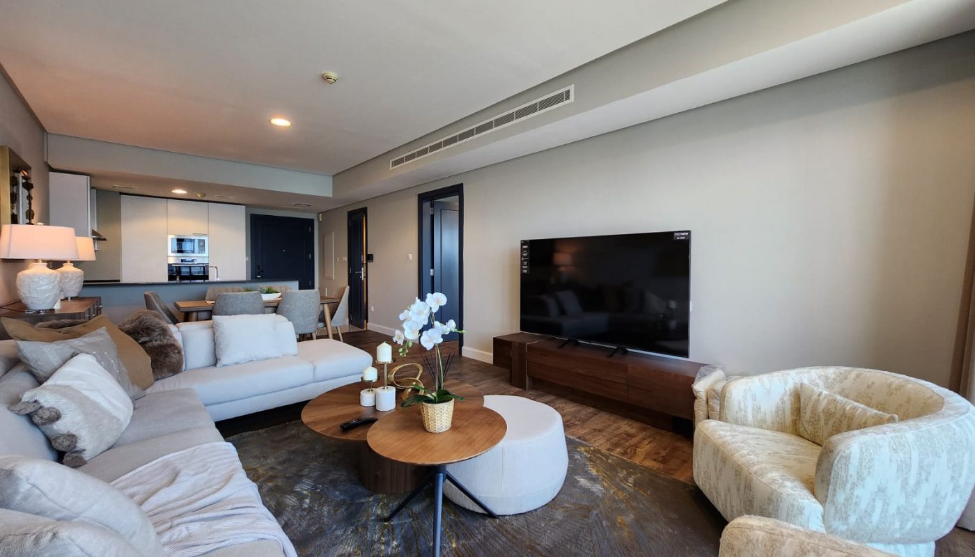 Modern living room with a large sectional sofa, round wooden coffee tables, a flat-screen TV, and neutral-colored decor. Kitchen area is visible in the background.