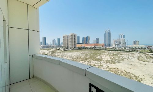 View from a luxury apartment balcony in the Sanabis area, showing a sandy landscape and city skyline with modern high-rise buildings under a clear blue sky.