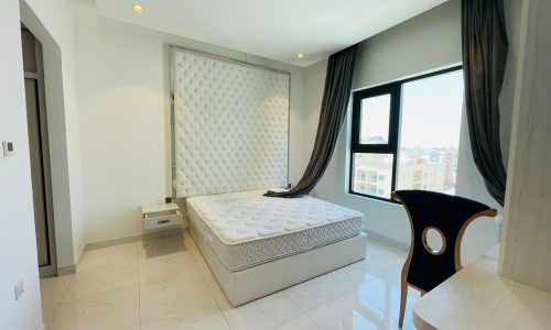 Modern bedroom with a large tufted headboard, king-sized bed, sleek black chair, and expansive window with curtains.