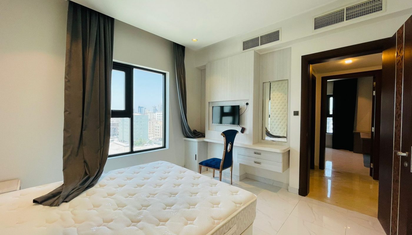 Modern bedroom with a large bed, desk and chair near a window, built-in tv and wardrobe, and an open door leading to another room.