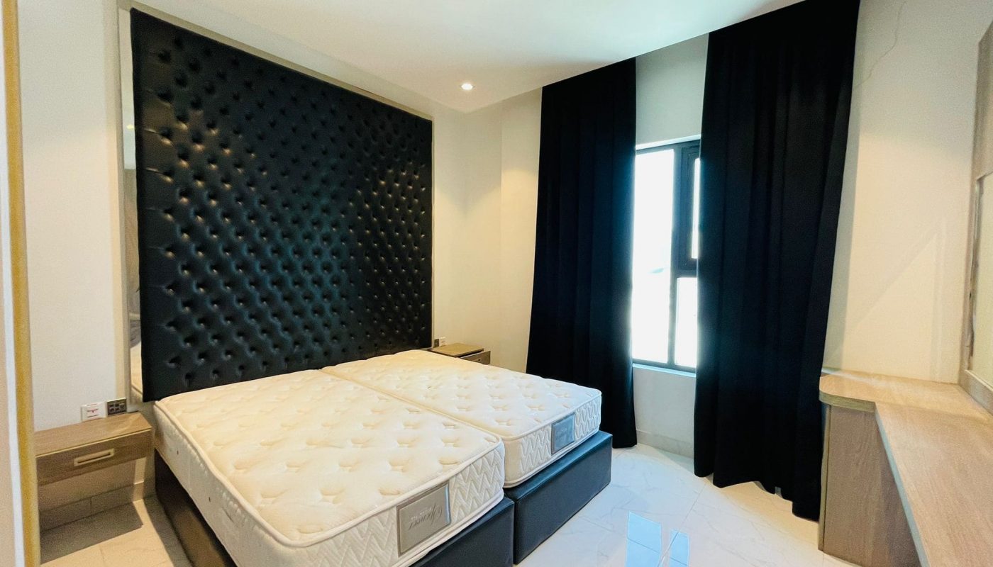 Modern bedroom with a large tufted headboard, two mattresses on a platform bed, black curtains, and a bright window.